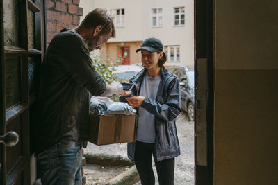 Side view of man doing digital signature on smart phone while receiving parcel from delivery person standing at doorway