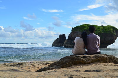 Rear view of couple sitting on rock by sea against sky