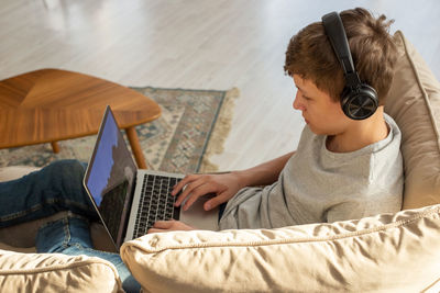 A teenage , sits on a sofa in a room, wearing black headphones , plays a computer game on a laptop.