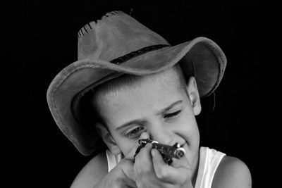 Close-up of boy wearing hat holding toy gun against black background