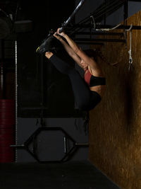 Side view of determined young fit sportswoman with long dark hair in activewear doing abs exercise hanging on metal bar while working out in modern gym