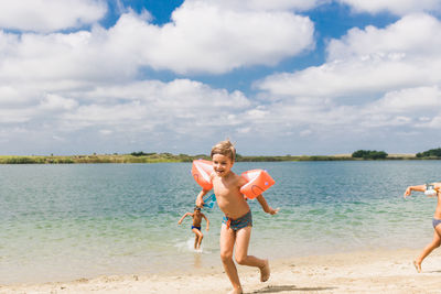 Full length of shirtless boy wearing water wings running on beach against cloudy sky