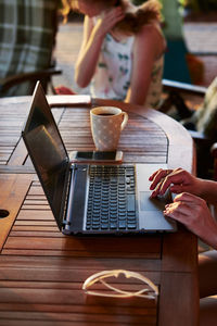 Cropped hands of girl using laptop by coffee cup on table