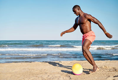 Full body shirtless african american male in shirts smiling and kicking colorful ball while having fun on sandy beach near waving sea on cloudless summer day