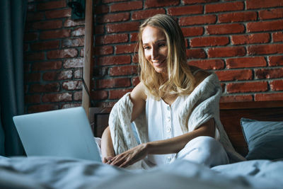 Adult woman forty years freelancer with blonde long hair in casual using laptop on bed at home