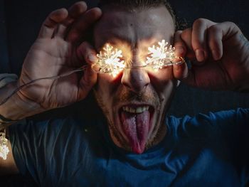 Close-up of aggressive man holding illuminated lighting equipment while sticking out tongue