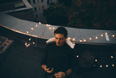 High angle view of young man using smart phone while relaxing on illuminated terrace at dusk