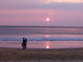Couple walking at beach against sky during sunset