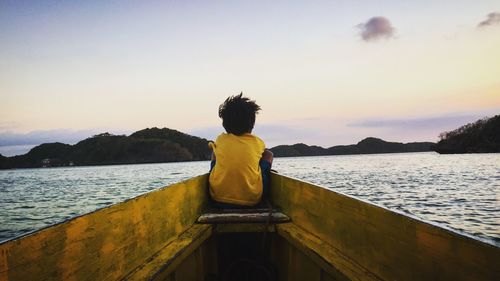 Rear view of boy looking at mountain while sitting on boat in lake against sky during sunset