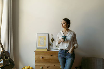 Thoughtful woman with cup standing and looking away at home