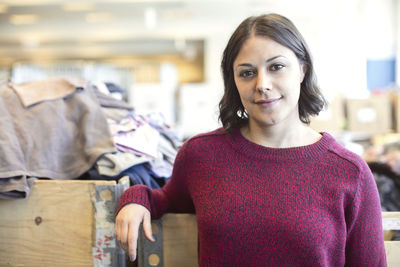 Portrait of smiling female volunteer standing against heap of clothes at workshop