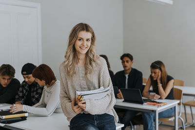 Portrait of happy young blond female student with book leaning on desk in classroom