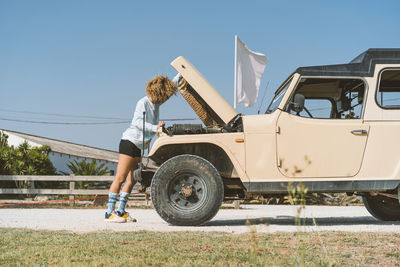 Young woman examining off-road vehicle while standing with open hood on sunny day