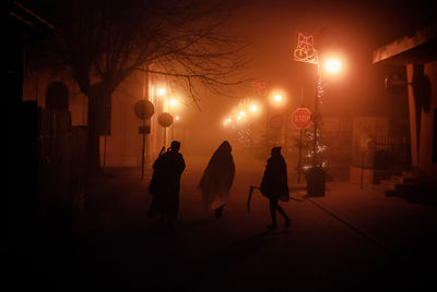 Silhouette people on road amidst illuminated lights during foggy weather at night