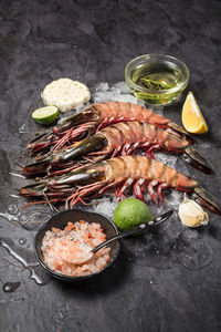 Three raw jumbo shrimps on ice with basic ingredients to cook, on dark background. vertical