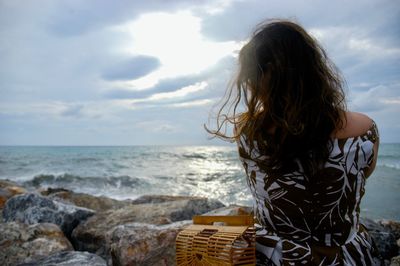 Rear view of woman sitting at beach against cloudy sky