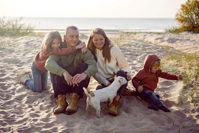 Family with a son and daughter and a dog sit on the sand in autumn