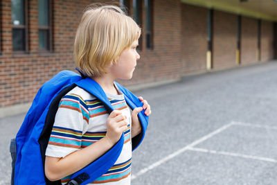 Beautiful child near school building at the schoolyard. little blond student with blue backpack. 