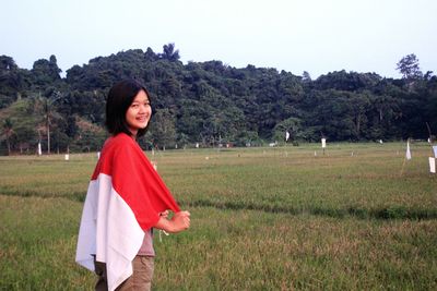 Smiling young woman holding monaco flag while standing on grass against sky