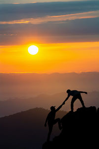 Silhouette man assisting friend to climb rock against sky during sunset