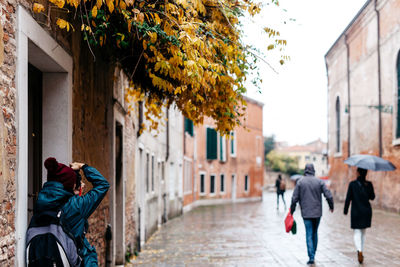 Rear view of person photographing people walking on street by building during rainy season