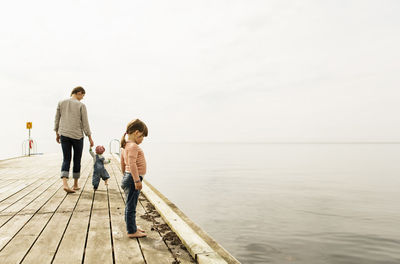 Side view of girl looking at sea while standing on pier with family walking in background against sky