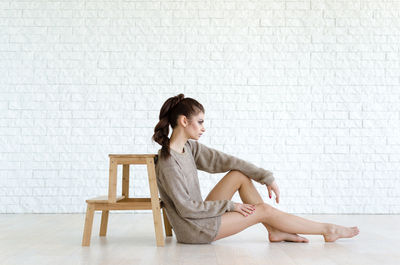 Full length of young woman sitting by wood stool against brick wall