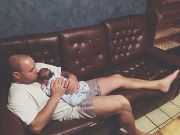 High angle view of father holding newborn baby while reclining on sofa at home