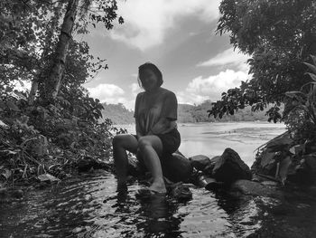 Full length of woman sitting on rock by trees against sky