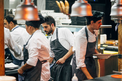 Men in hats and aprons working in team and serving dishes with delicious food in kitchen