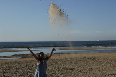 Playful girl throwing sand at beach against sky