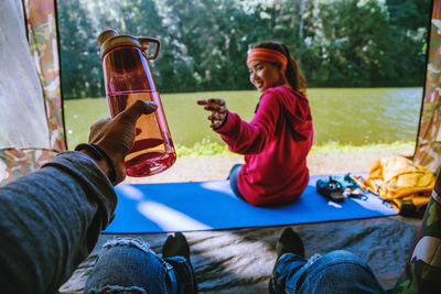 Midsection of man holding bottle with woman sitting in background