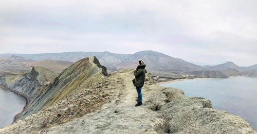 Side view of young man standing on mountain by lake against sky