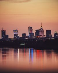 Illuminated buildings by river against sky during sunset