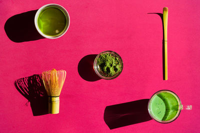 Top view of bright background of glass of green tea and dried matcha with bamboo chasen and chashaku on pink surface