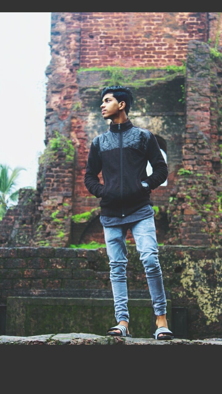 one person, casual clothing, young men, young adult, day, real people, leisure activity, architecture, full length, wall, lifestyles, standing, nature, built structure, outdoors, front view, looking, building exterior, looking away, brick, jeans, teenager, contemplation