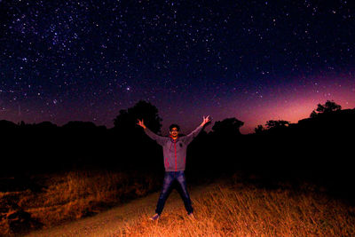 Full length of young man with arms outstretched standing on field against star field at night
