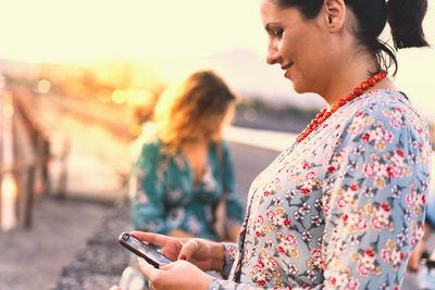 Side view of mature woman using smart phone while standing against sky during sunset