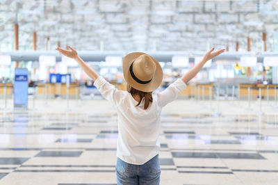 Rear view of woman wearing hat with arms raised standing at airport