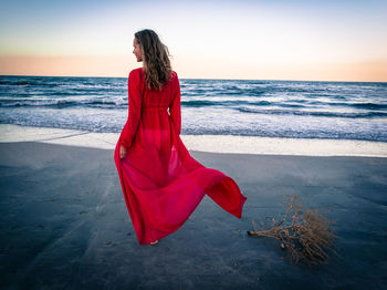 Woman in red dress walking at beach against sky during sunset