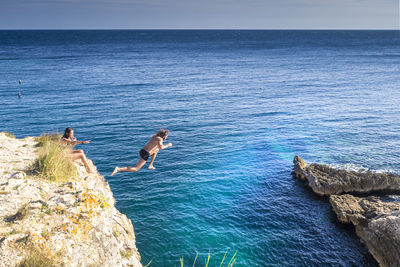 Man jumping by woman on rocks over sea against sky