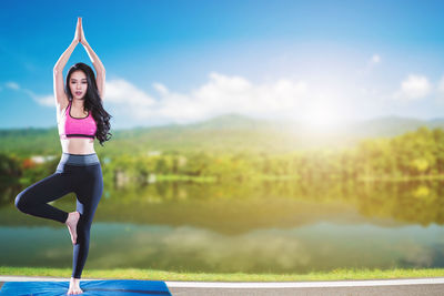 Full length portrait of beautiful woman doing tree pose by lake against sky