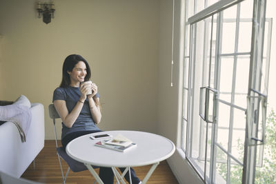 Smiling thoughtful woman having coffee while looking through window at table in living room