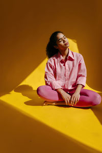 Young woman with eyes closed sitting cross-legged against yellow background