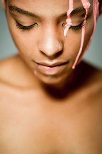 Creative ethnic female model with pink paint dripping on her face looking down on gray background in studio