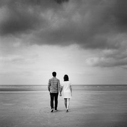 Rear view of couple walking at beach against cloudy sky