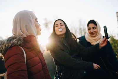 Multi-ethnic female friends blowing bubble gums while taking selfie against sky