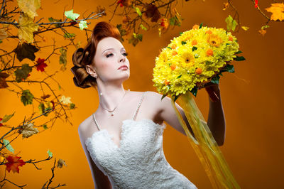 Beautiful bride looking at bouquet while standing against orange background