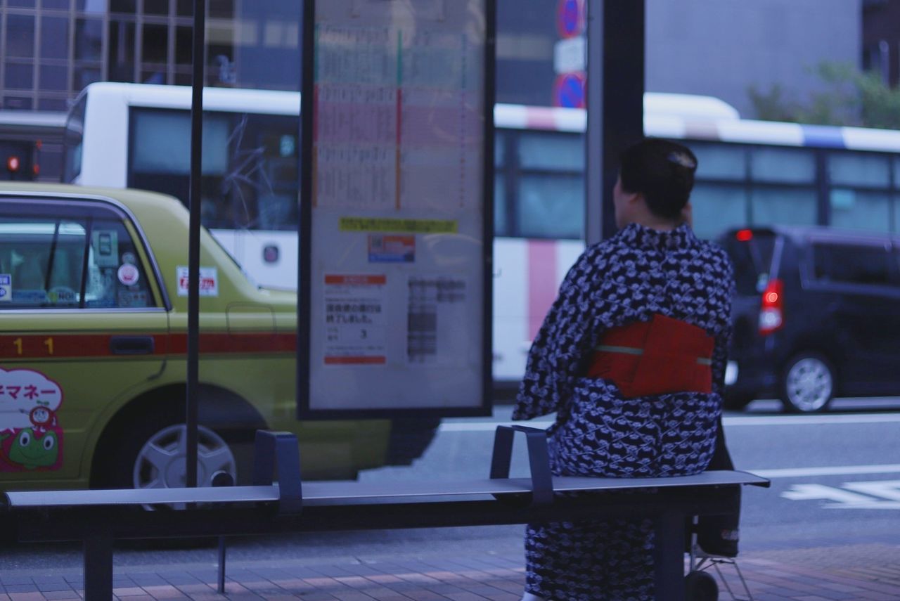 mode of transportation, transportation, one person, real people, land vehicle, rear view, public transportation, bus, lifestyles, city, women, leisure activity, street, architecture, incidental people, adult, motor vehicle, day, standing, outdoors, warm clothing, waiting