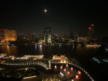 Illuminated modern buildings by river against sky at night